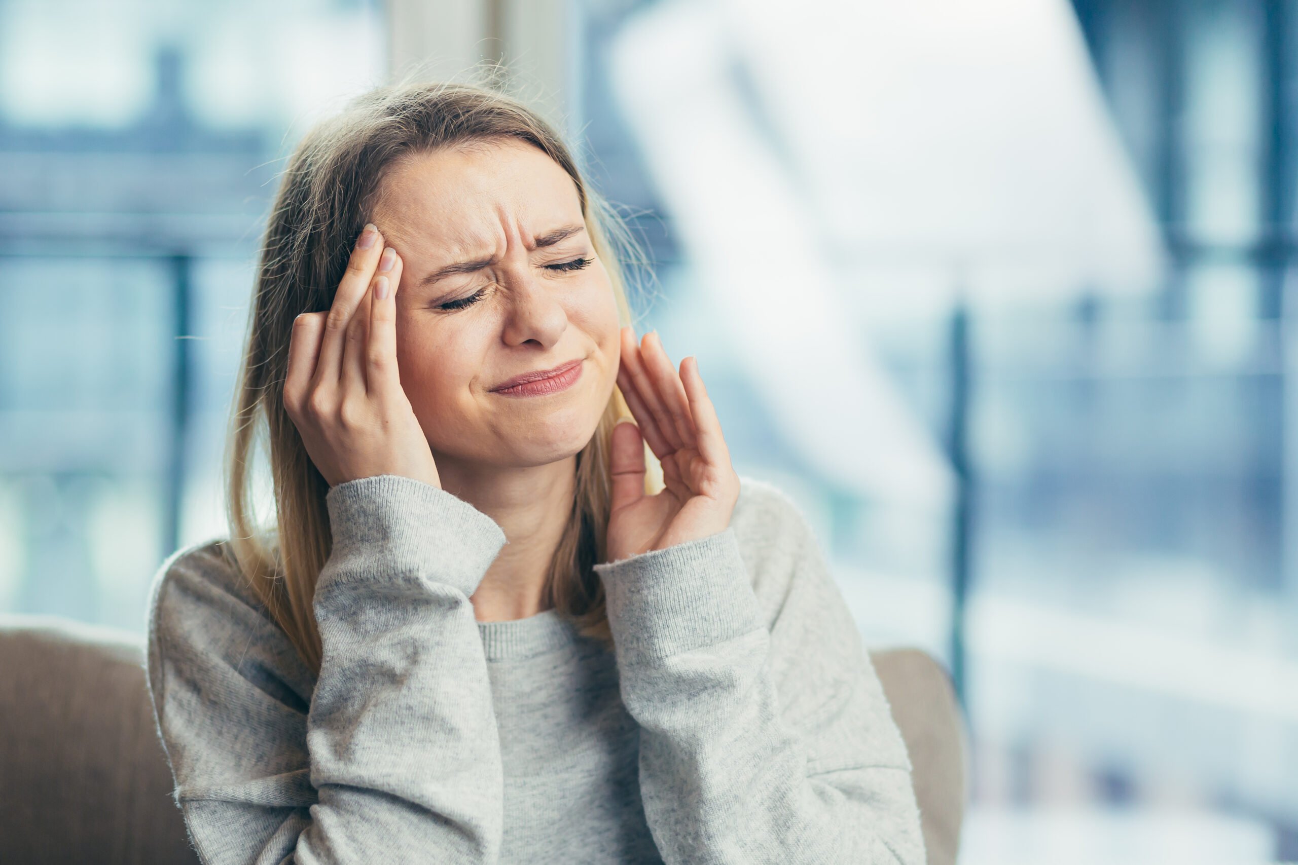 A woman in a grey jumper sits indoors, eyes closed and face tense, pressing her fingers to her temple and jaw as if experiencing pain or discomfort—perhaps from Invisalign-related headaches.