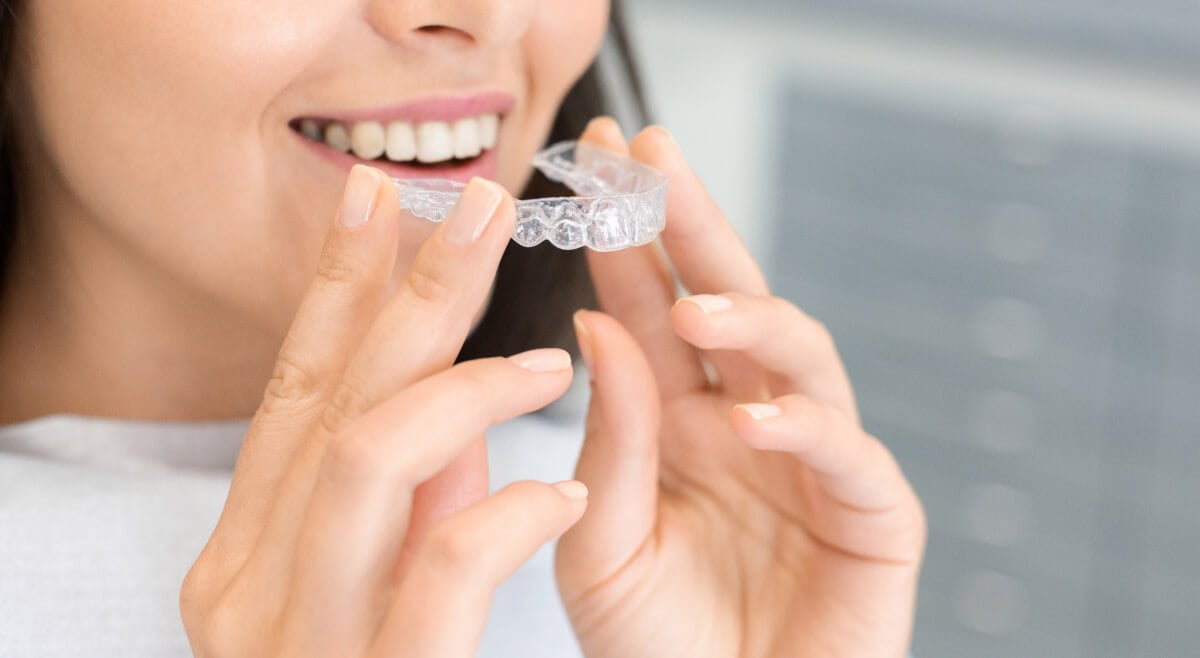 A person smiling whilst holding and about to put on a clear dental aligner, with focus on their hands, teeth, and the aligner—perfectly capturing the Invisalign experience for those seeking Invisalign information.