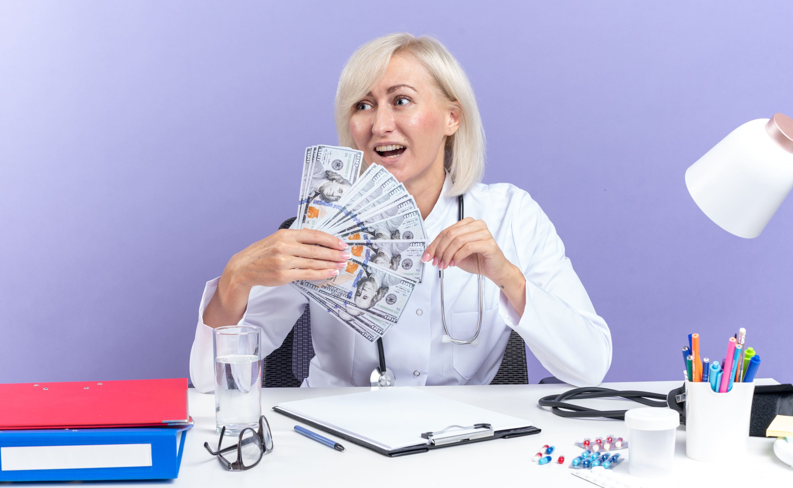 A doctor in a white coat sits at a desk, smiling and holding a fan of hundred-dollar notes. Medical files, a stethoscope, pills, and a glass of water are on the desk, alongside an image from an Ipswich dentist CT scanner, all set against a purple background.