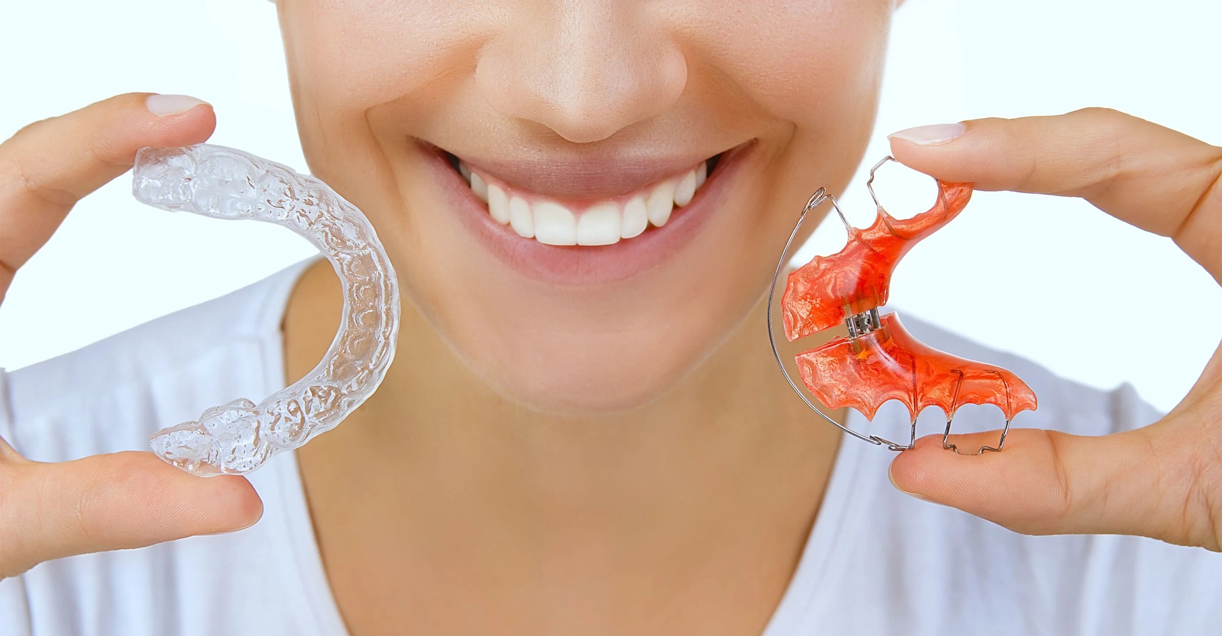 A person smiles while holding a clear dental aligner in one hand and a red orthodontic retainer in the other hand, showing a comparison between the two dental devices and highlighting the importance of wearing a retainer after Invisalign treatment.