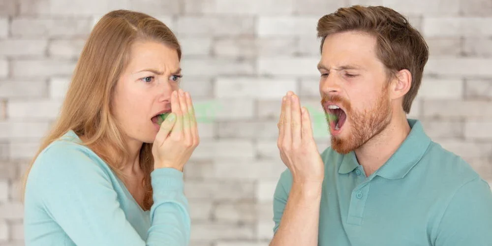 A woman and a man in light blue shirts check their breath by exhaling into their hands and smelling it, standing in front of a light brick wall, perhaps wondering: Does composite bonding cause bad breath?.