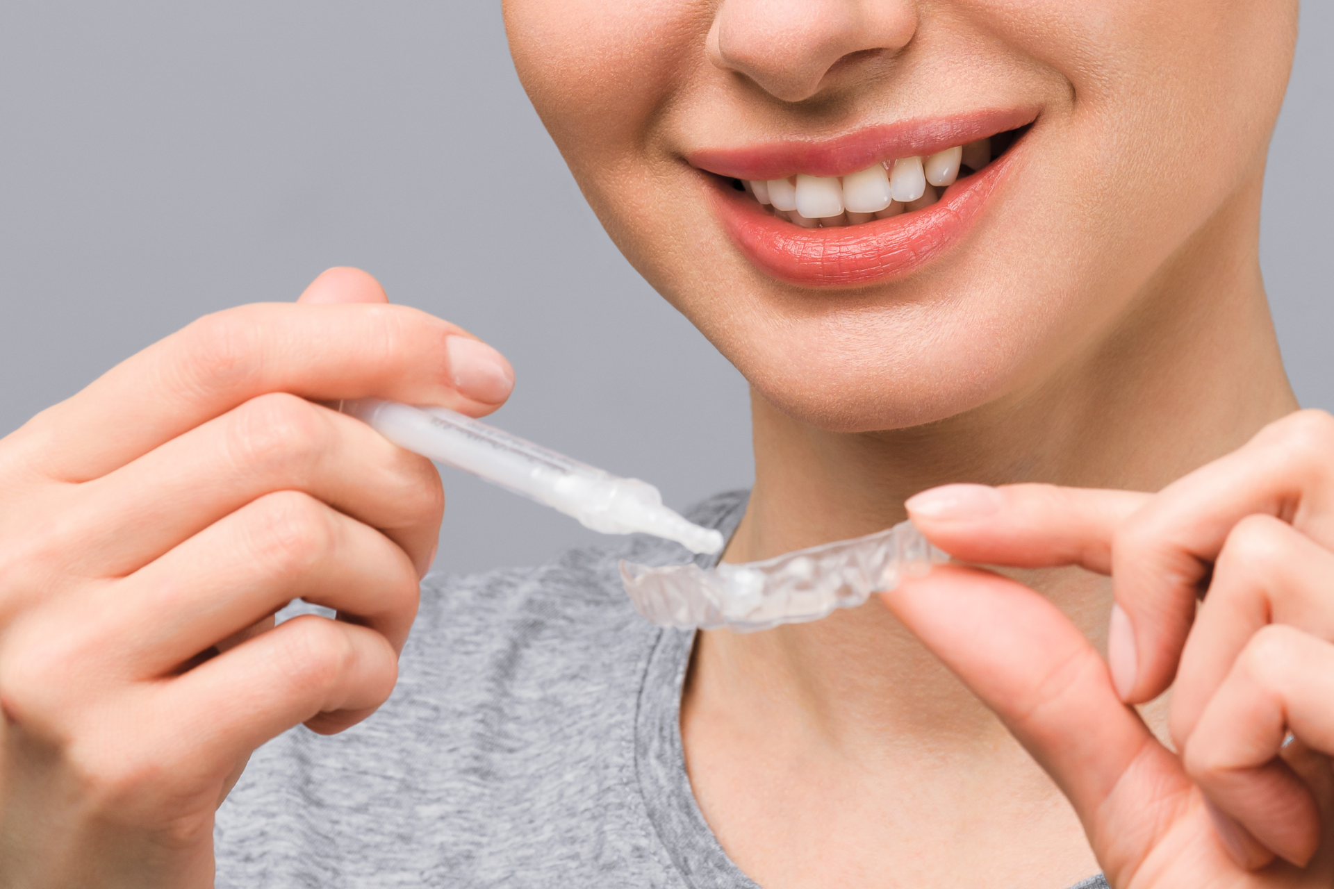A woman smiling whilst applying gel from a syringe onto a clear dental aligner, preparing it for use. She is wearing a grey shirt and the background is plain and light-coloured. Similar to popular teeth whitening pens, this simple at-home method aims to brighten her smile with ease and convenience.