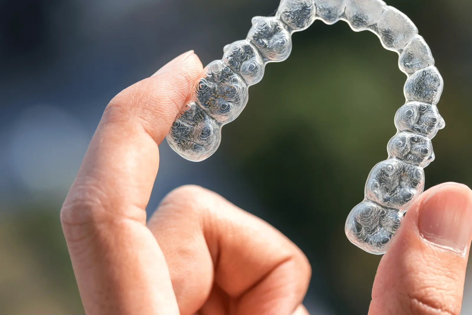 A hand holds a clear dental aligner, commonly used for teeth straightening and addressing issues such as crossbite, against a blurred outdoor background, prompting the question: Can Invisalign fix crossbite?.