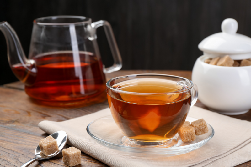 A glass cup of tea on a saucer with brown sugar cubes and a spoon nearby, perfect for enjoying tea with Invisalign. In the background, a glass teapot filled with more tea and a sugar bowl sit on a wooden table, creating an inviting scene for those who love drinking tea even whilst wearing Invisalign.