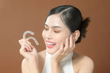A smiling woman holds a clear Invisalign retainer in one hand while resting her chin on the other, looking thoughtfully at the aligner as she considers how often to wear it against a brown background.